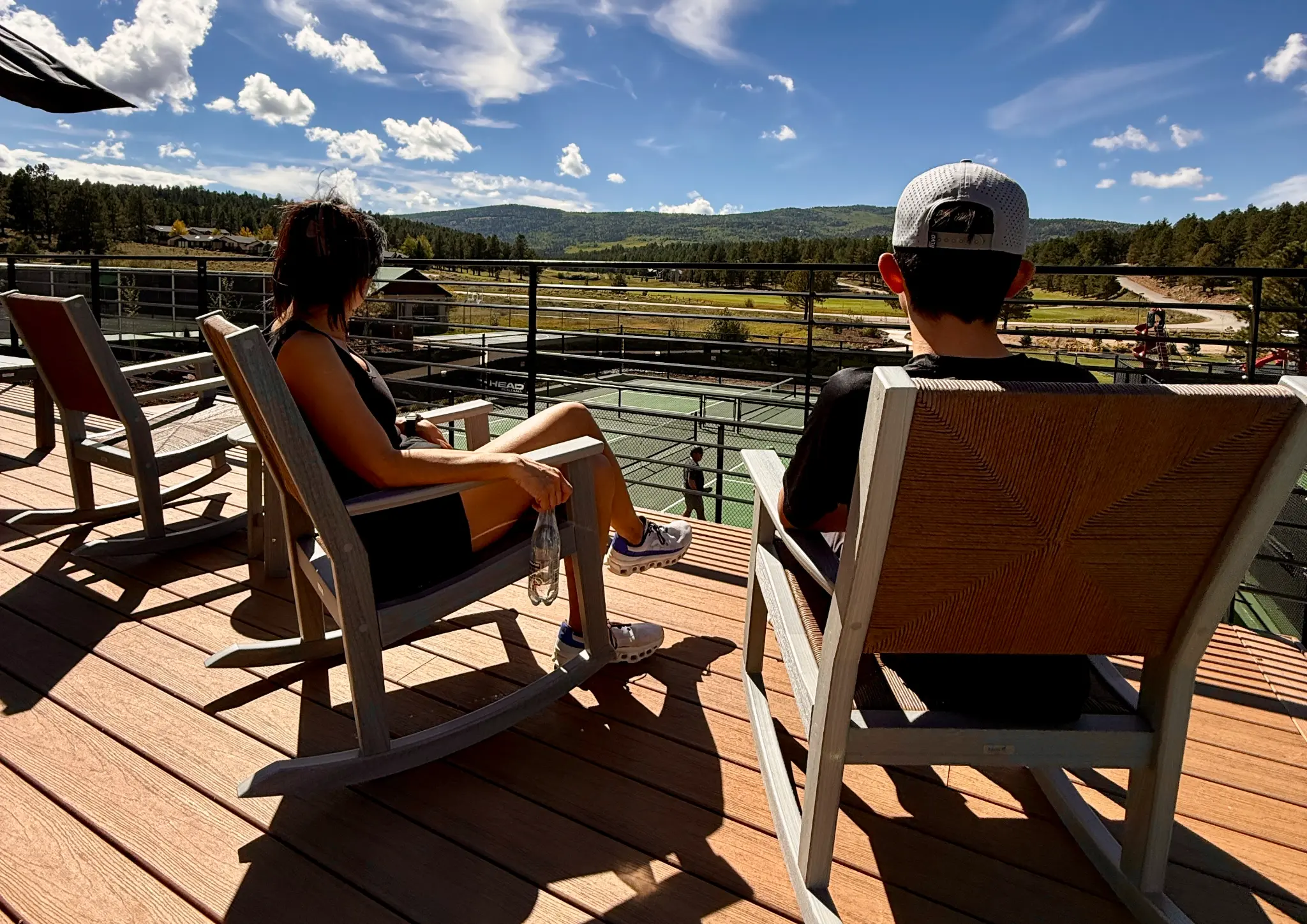 Family relaxing in rocking chairs on the upper deck above Angel Fire Resort's Olympic Park pickleball courts with mountain views